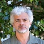 Headshot of a person with short grey hair and beard standing in front of greenery