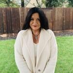 Picture of a slightly smiling person with long, dark hair and red lipstick wearing a white jacket standing on a lawn.