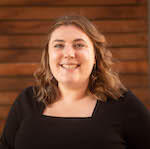 Headshot of a smiling person with medium-length brown hair and a dark top in front of a wood wall.