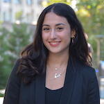 Headshot of smiling person with long dark hair wearing a dark top and necklace in front of a building and greenery.