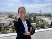 Headshot of a smiling person with blond hair pulled back, wearing a dark blazer over a white top, crossing their arms in front of a view of campus in the background