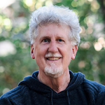 Headshot of a smiling person with short grey hair and goatee in front of greenery