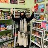 A masked person with long dark hair and a scarf standing with hands raised in front of shelves of food in a food pantry. 