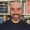 Headshot of a smiling person with shaved head and beard in front of a bookshelf.