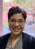 Headshot of a smiling person with short dark reddish hair, wearing glasses and hoop earrings.