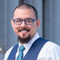 Headshot of a smiling person with short dark hair and facial hair wearing glasses and a collared shirt,