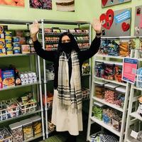 A person with long dark hair and a mask raising her hands in the Cal NERDS food pantry, in front of well-stocked shelves.