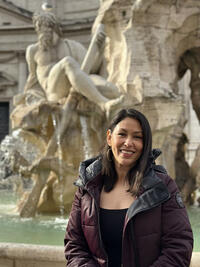 Photo of a smiling person with shoulder-length dark hair from the waist up in front of a fountain with sculptures.