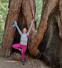 Photo of a smiling person in tree pose standing in front of old growth redwoods.