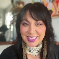 Headshot of a smiling person with long dark hair and bangs wearing a scarf and long feather earrings.