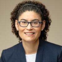 Headshot of a smiling person with medium-length curly hair wearing glasses and a headband.