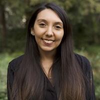 Headshot of a smiling person with long dark hair in front of greenery.