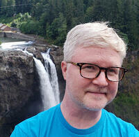 Headshot of a smiling person with short light hair and glasses and a blue t-shirt, in front of a waterfall with lush green trees in the background.