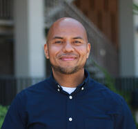 Headshot of smiling person with a shaved head and neatly trimmed facial hair wearing a dark button down shirt with collar.