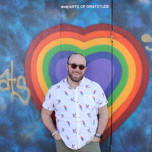 Photo of a smiling person with sunglasses and beard posing with their hands in their pockets in front of a wall painted with a rainbow heart.